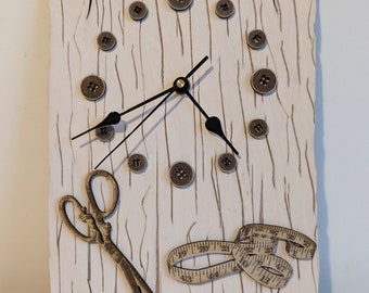 May include: A white wooden clock with a black clock face and hands. The clock is decorated with brown buttons and a gold measuring tape and scissors.
