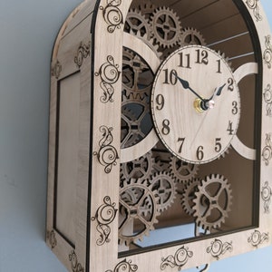May include: A wooden clock with a decorative, ornate frame and a pendulum. The clock face has Roman numerals and a set of gears visible behind it.