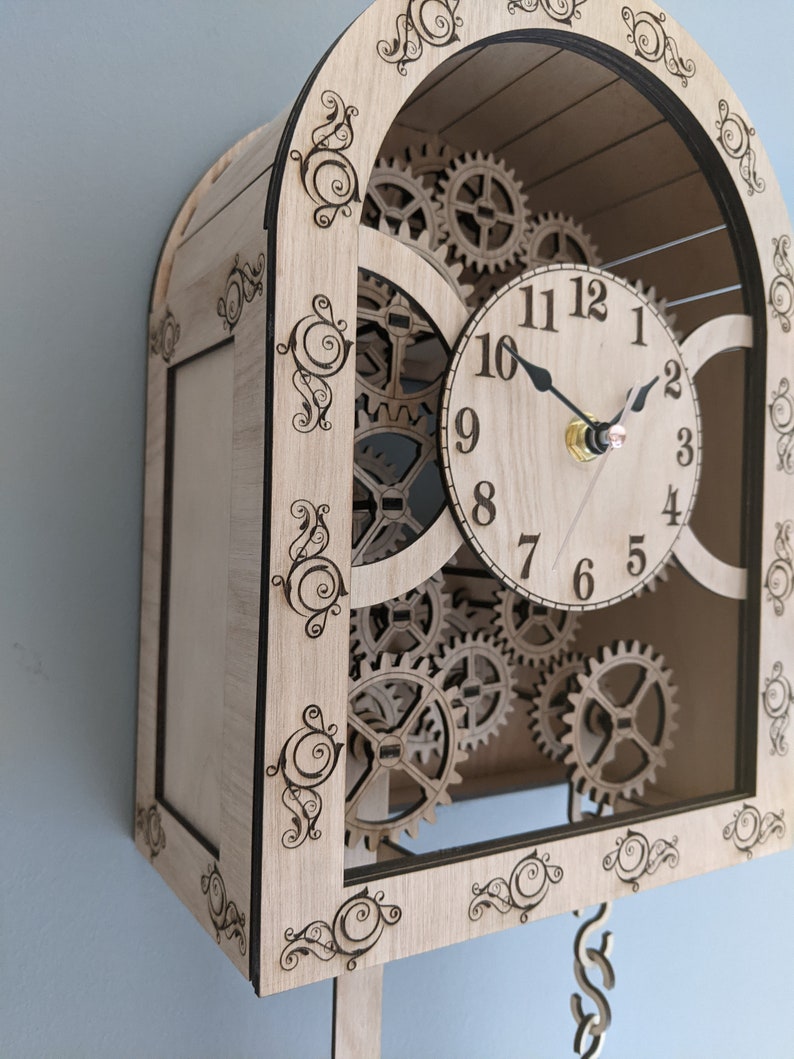May include: A wooden clock with a decorative, ornate frame and a pendulum. The clock face has Roman numerals and a set of gears visible behind it.