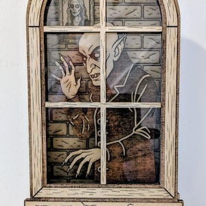 Puede incluir: Decoración de madera con temática de Nosferatu. El diseño presenta una figura de vampiro mirando a través de una ventana con un fondo de ladrillos y un retrato enmarcado. La palabra "NOSFERATU" se muestra en la base.
