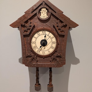 Op de afbeelding: Een bruine houten koekoeksklok met een decoratief ontwerp. De ronde wijzerplaat toont Romeinse cijfers. De klok heeft een klein rond venster aan de bovenkant met een gebeeldhouwde figuur. De klok heeft kettingachtige pendels.