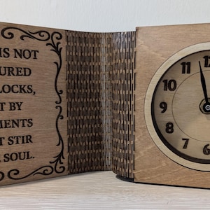 Op de afbeelding: Houten klok met een lichtbruine afwerking. De wijzerplaat heeft zwarte cijfers en wijzers. De linkerkant van de klok heeft de quote: "TIJD WORDT NIET GEMETEN DOOR KLOKKEN, MAAR DOOR MOMENTEN DIE DE ZIEL ROEREN."