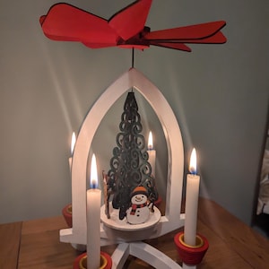 Peut inclure: Une pyramide de Noël en bois blanc avec une hélice rouge sur le dessus. La pyramide a quatre bougeoirs avec des bougies blanches. Le centre de la pyramide présente un sapin de Noël vert découpé au laser et un bonhomme de neige.