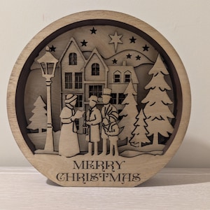 Può includere: Decorazione natalizia rotonda in legno con un design a strati. La scena raffigura un gruppo di cantori di canti natalizi davanti a case, un lampione e alberi di Natale. Le parole "MERRY CHRISTMAS" sono incise in basso. Stelle e una stella filante sono scolpite sullo sfondo.