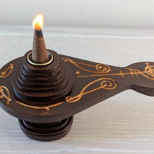 May include: A brown wooden incense cone holder with gold-coloured etched designs. A lit incense cone sits in the centre, emitting a flame. The holder has a tiered design and a long, flat handle.