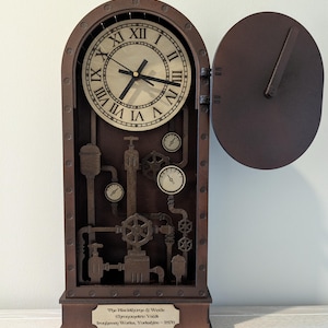 May include: A steampunk-style clock with a dark brown wooden frame and a light-colored clock face featuring Roman numerals. The clock has an open side door and decorative gauges and pipes. The text on the bottom reads "The Blackdhorpe & Wede Chronometric Vault Ironjaven Works, Yorkshire - 1876."