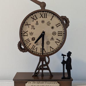Pode incluir: Um relógio steampunk castanho com algarismos romanos e símbolos celestes. Possui um braço decorativo e uma pequena figura de uma pessoa com um telescópio. A base diz "Dr. Archibald Fenwick's Celestial Regulator ~ Est. 1871 ~."