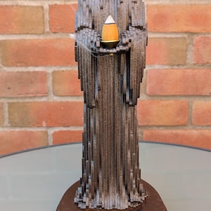 Pode incluir: Uma escultura de figura encapuzada, cinzento-escura e em camadas, sobre uma base circular de madeira. A figura segura um pequeno prato cor de latão com um incenso cónico aceso que emite um fino rasto de fumo pelo topo.