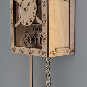 May include: A wooden clock with a pendulum and intricate gears visible through the front. The clock face has Roman numerals and black hands. The clock is made of light wood with dark brown accents.