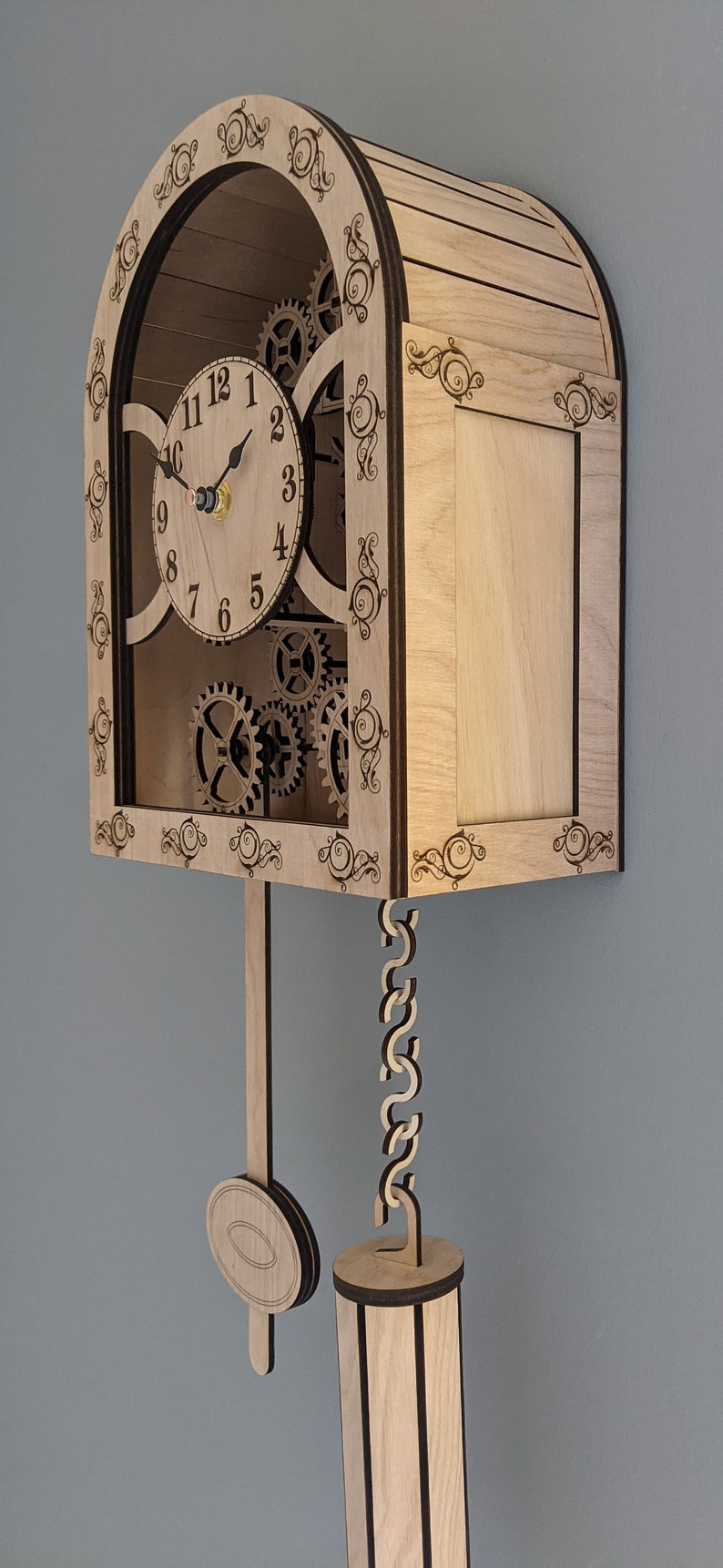 May include: A wooden clock with a pendulum and intricate gears visible through the front. The clock face has Roman numerals and black hands. The clock is made of light wood with dark brown accents.