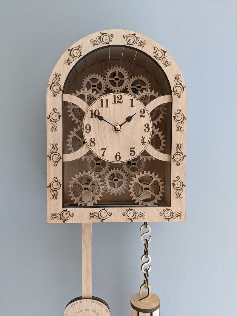 May include: A wooden clock with a pendulum and intricate gears visible behind the clock face. The clock face has Roman numerals and black hands.