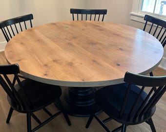 May include: A round wooden dining table with a light brown surface and a dark base, surrounded by four black chairs. The chairs have a spindle back design. The table is set on a hardwood floor.