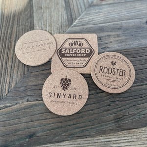 May include: A collection of cork coasters with various logos and text. One coaster reads "SEEDS & SAWDUST", another "SALFORD COFFEE SHOP", a third "GINYARD premium brewery", and the last "ROOSTER BREWING & CO". The coasters are round and square.
