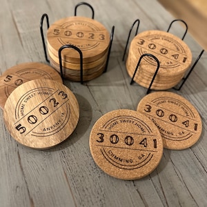 May include: Set of six wooden coasters with a cork top. Each coaster features a circular design with the text "Home Sweet Home" and a unique number. The coasters are arranged in two stacks, one on a black metal stand and the other on a wooden table.