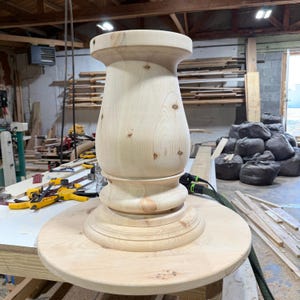 May include: A light-colored, unfinished wooden table pedestal base featuring a smooth, curved, hourglass-shaped central column mounted onto a circular flat tabletop surface, showcasing natural wood grain patterns and several small knots.