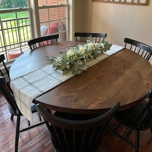 May include: A round dark brown wooden dining table surrounded by six black spindle-back chairs. A rectangular table runner with a white and black plaid pattern lies across the center, topped with a centerpiece of green foliage and candles.