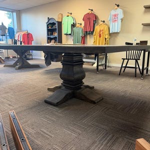 May include: A large, dark-stained wooden table with a thick, rectangular top and a central pedestal base featuring a decorative, tiered, and curved design. The table stands on a wide, four-pronged base, showcasing a visible wood grain texture.