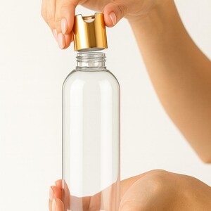 K&ouml;nnte beinhalten: Eine transparente, zylindrische Flasche mit einem goldfarbenen Klappdeckel. Die Flasche wird von zwei H&auml;nden vor einem wei&szlig;en Hintergrund gehalten. Der Deckel ist ge&ouml;ffnet und gibt die Flaschen&ouml;ffnung frei.
