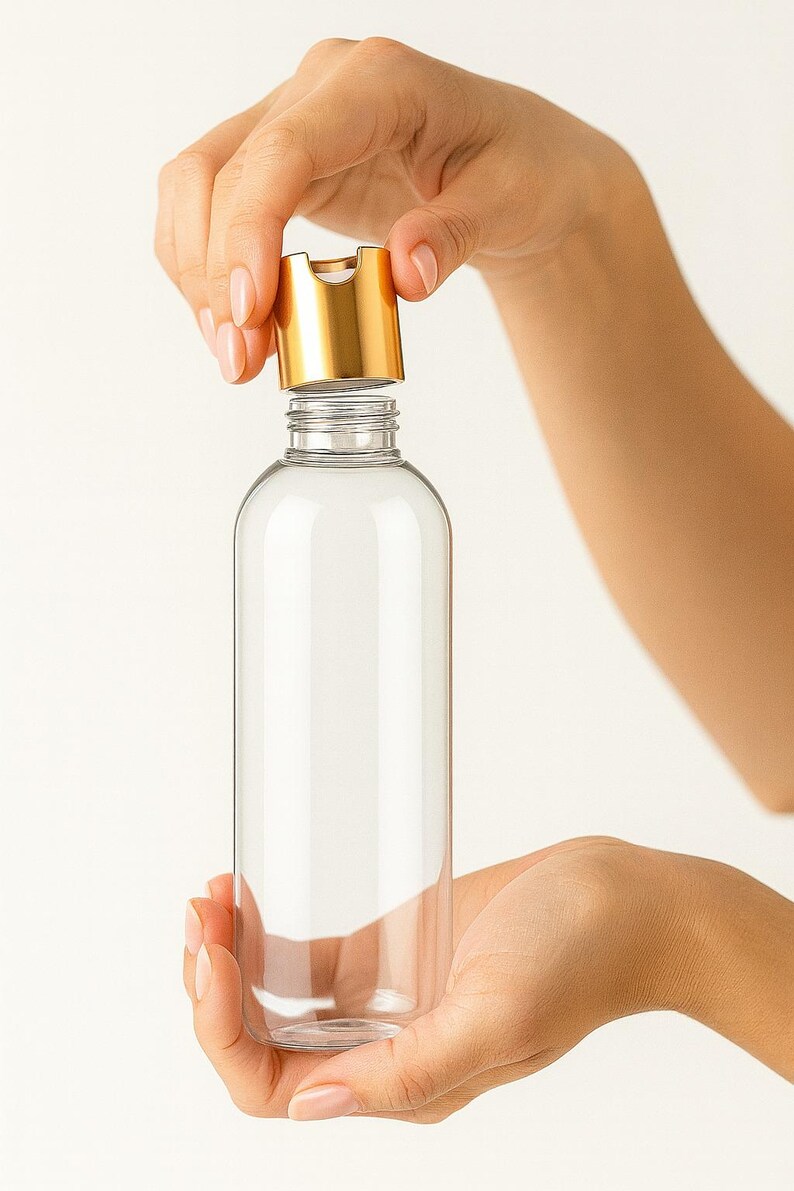 K&ouml;nnte beinhalten: Eine transparente, zylindrische Flasche mit einem goldfarbenen Klappdeckel. Die Flasche wird von zwei H&auml;nden vor einem wei&szlig;en Hintergrund gehalten. Der Deckel ist ge&ouml;ffnet und gibt die Flaschen&ouml;ffnung frei.
