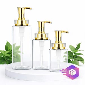 May include: Three clear, rectangular soap or lotion dispensers with gold-colored pump tops. The dispensers are arranged in a row, decreasing in size from left to right. The background is white with green foliage on the sides.