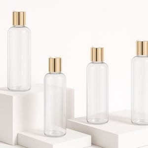 K&ouml;nnte beinhalten: Vier transparente, zylindrische Flaschen mit goldfarbenen Verschl&uuml;ssen werden auf wei&szlig;en geometrischen Plattformen pr&auml;sentiert. Die Flaschen sind leer, was darauf hindeutet, dass sie f&uuml;r Kosmetik- oder K&ouml;rperpflegeprodukte bestimmt sind. Das minimalistische Design und die neutrale Farbpalette schaffen eine saubere, moderne &Auml;sthetik.