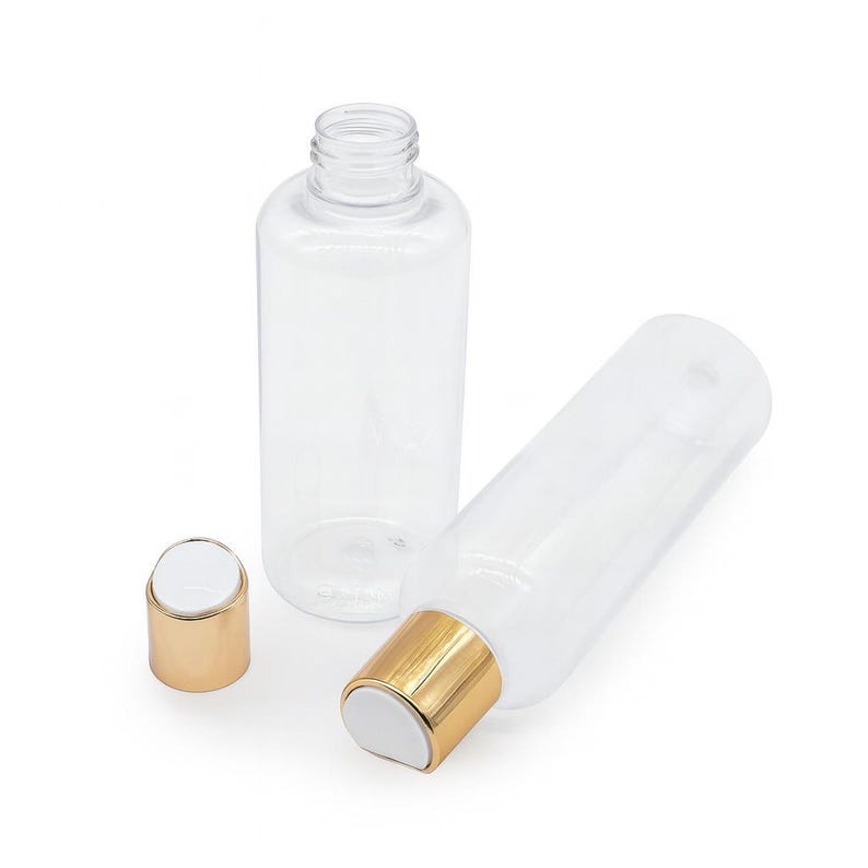 K&ouml;nnte beinhalten: Transparente Kunststoffflaschen mit goldfarbenen Verschl&uuml;ssen. Eine Flasche steht aufrecht, eine andere liegt auf der Seite, und ein dritter Verschluss ist separat. Die Flaschen sind zylindrisch und scheinen leer zu sein.