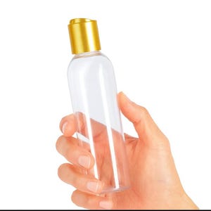 K&ouml;nnte beinhalten: Eine transparente, zylindrische Flasche mit einem goldfarbenen Deckel. Die Flasche wird in einer Hand vor einem wei&szlig;en Hintergrund gehalten. Die Flasche ist leer und scheint aus Kunststoff zu sein.