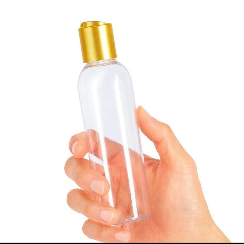 K&ouml;nnte beinhalten: Eine transparente, zylindrische Flasche mit einem goldfarbenen Deckel. Die Flasche wird in einer Hand vor einem wei&szlig;en Hintergrund gehalten. Die Flasche ist leer und scheint aus Kunststoff zu sein.