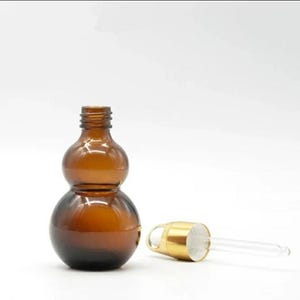 May include: A brown glass dropper bottle with a gourd-like shape. The bottle has a gold-colored cap and a clear glass dropper. The bottle is empty and sits on a white background. The bottle is likely used for essential oils or serums.