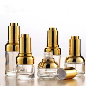 May include: Clear glass dropper bottles with gold-coloured caps. The bottles are empty, with a pipette inside. One bottle has its cap removed, showing the dropper. The bottles are arranged on a light-coloured surface.