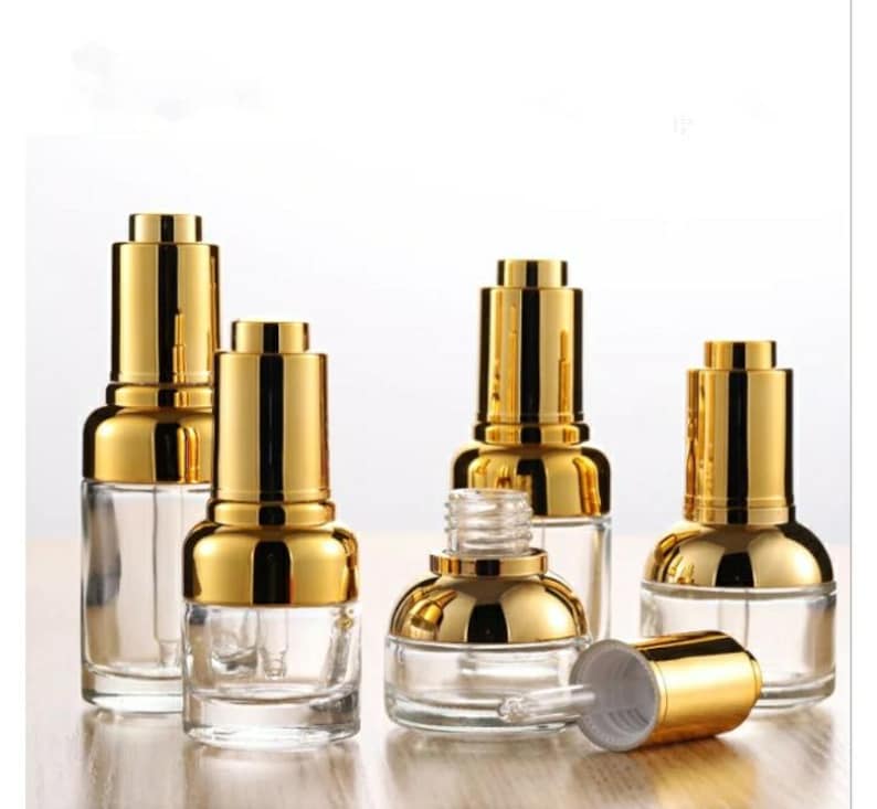 May include: Clear glass dropper bottles with gold-coloured caps. The bottles are empty, with a pipette inside. One bottle has its cap removed, showing the dropper. The bottles are arranged on a light-coloured surface.