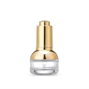 May include: A clear glass dropper bottle with a gold-coloured cap and top. The bottle has a rounded base and a long, cylindrical gold cap. The dropper is visible inside the bottle. Likely for cosmetics or skincare products.