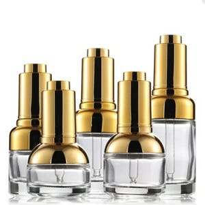 May include: Several clear glass dropper bottles with gold-coloured caps. The bottles have a rounded base and a long, thin dropper inside. The gold caps have a shiny, metallic finish. The bottles are arranged in a group against a white background.