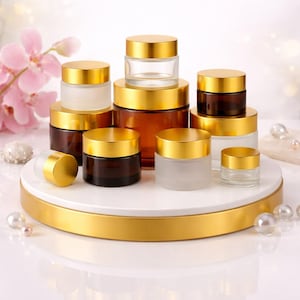 May include: An assortment of glass cosmetic jars with gold-colored lids. The jars vary in size and color, including clear, frosted, and amber. They are arranged on a white and gold display, with pearls and pink flowers in the background.