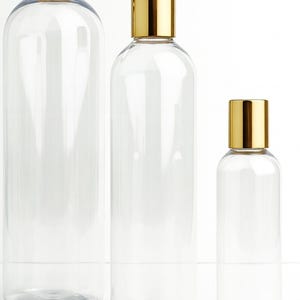 K&ouml;nnte beinhalten: Drei transparente, zylindrische Flaschen in verschiedenen Gr&ouml;&szlig;en mit goldfarbenen Deckeln. Die Flaschen sind leer und haben ein schlichtes, modernes Design. Die Deckel sind rechteckig und b&uuml;ndig mit der &Ouml;ffnung der Flasche. Die Flaschen sind vertikal vor wei&szlig;em Hintergrund angeordnet.