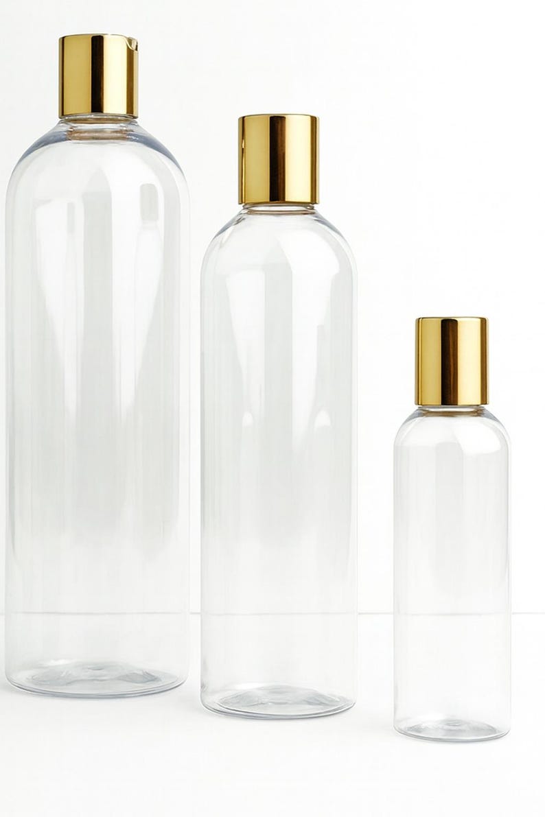 K&ouml;nnte beinhalten: Drei transparente, zylindrische Flaschen in verschiedenen Gr&ouml;&szlig;en mit goldfarbenen Deckeln. Die Flaschen sind leer und haben ein schlichtes, modernes Design. Die Deckel sind rechteckig und b&uuml;ndig mit der &Ouml;ffnung der Flasche. Die Flaschen sind vertikal vor wei&szlig;em Hintergrund angeordnet.