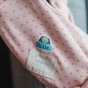 May include: Pink baby onesie with a small star pattern. A personalized name tag with a turtle design reads "Ellie". The onesie has a white tag that reads "pehr indigobaby".