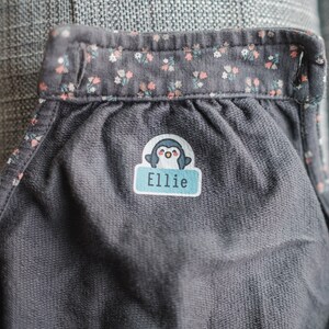 May include: A gray fabric with a floral print border. A blue and white penguin sticker with the name "Ellie" is attached to the fabric.