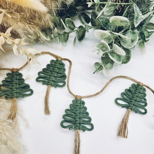 May include: A garland of four green crocheted Christmas trees with brown twine tassels hanging on a white background.