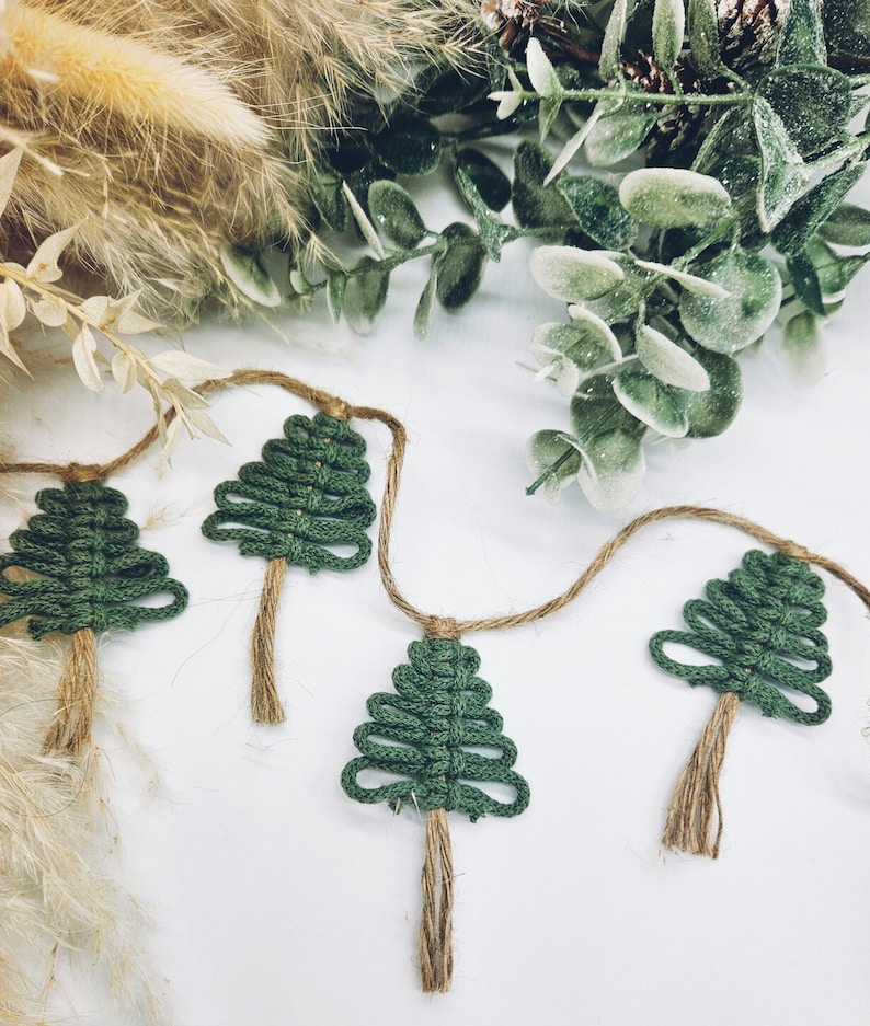 May include: A garland of four green crocheted Christmas trees with brown twine tassels hanging on a white background.