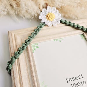 May include: A wooden picture frame with a green crocheted hanger and a white crocheted daisy with a yellow center. The frame has a white background with green leaves printed on it. The text "Insert Photo" is printed on the frame.