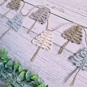 May include: A garland of crocheted trees in shades of beige, brown, and grey, strung together with twine. The garland is displayed on a white wooden surface with green foliage.