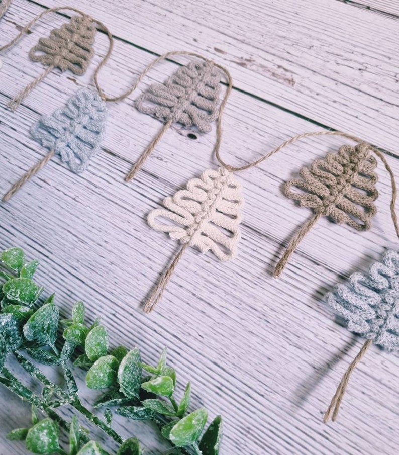 May include: A garland of crocheted trees in shades of beige, brown, and grey, strung together with twine. The garland is displayed on a white wooden surface with green foliage.