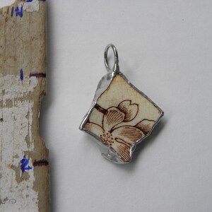 Peut inclure: Un pendentif en argent avec un motif floral brun sur fond blanc. Le pendentif est en forme de diamant et a une boucle en haut pour l'accrocher.