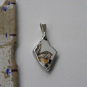 Op de afbeelding: Een zilveren hanger met een diamantvormig stuk wit porselein met een bruin en geel bloemenontwerp.