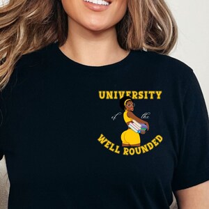 May include: Black t-shirt with a graphic that reads "UNIVERSITY of the WELL ROUNDED." The graphic features a cartoon woman in a yellow outfit holding books. The text and graphic are in yellow and white.