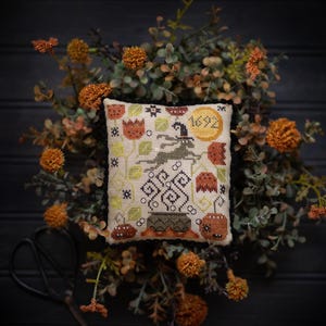May include: A square cross-stitch sampler titled Salem Mini Moon featuring a central witch riding a broomstick, a large yellow moon with the year 1692, a black cauldron, pumpkins, and floral motifs in shades of orange, green, and black on beige fabric.