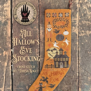 May include: A mustard-colored fabric stocking featuring a cross-stitch design of a haunted house, a pumpkin-headed figure, ghosts, bats, and a black cat. The text "All Hallow's Eve" is stitched near the bottom, and a braided loop is attached at the top.