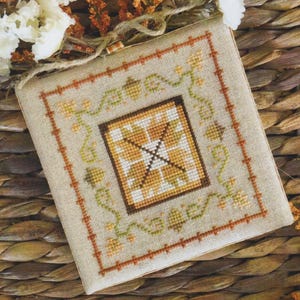 May include: A square fabric piece featuring a cross-stitch design on a light beige background. The centre displays a geometric star pattern in shades of yellow and brown, surrounded by a decorative border of green vines and orange cross-stitch lines.
