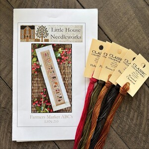 May include: A cross stitch pattern for a bookmark with the text "Farmers Market ABC's" and the product number "LHN-210". The pattern features a small house, a tree, and a basket of fruit. The pattern is accompanied by five skeins of embroidery floss in the colours red, green, brown, black, and orange.